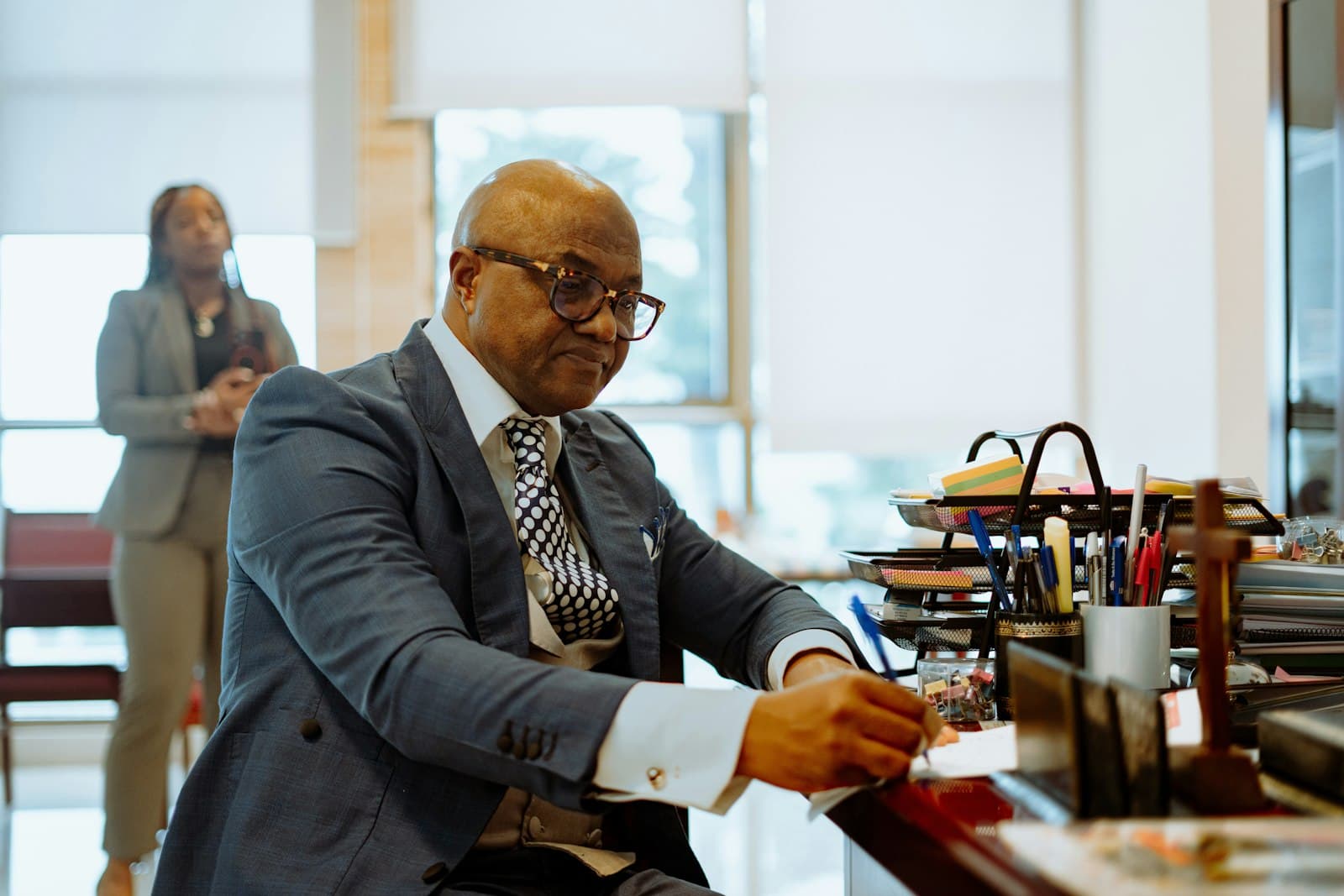 An attorney in a grey suit writing at his desk while a colleague stands behind him